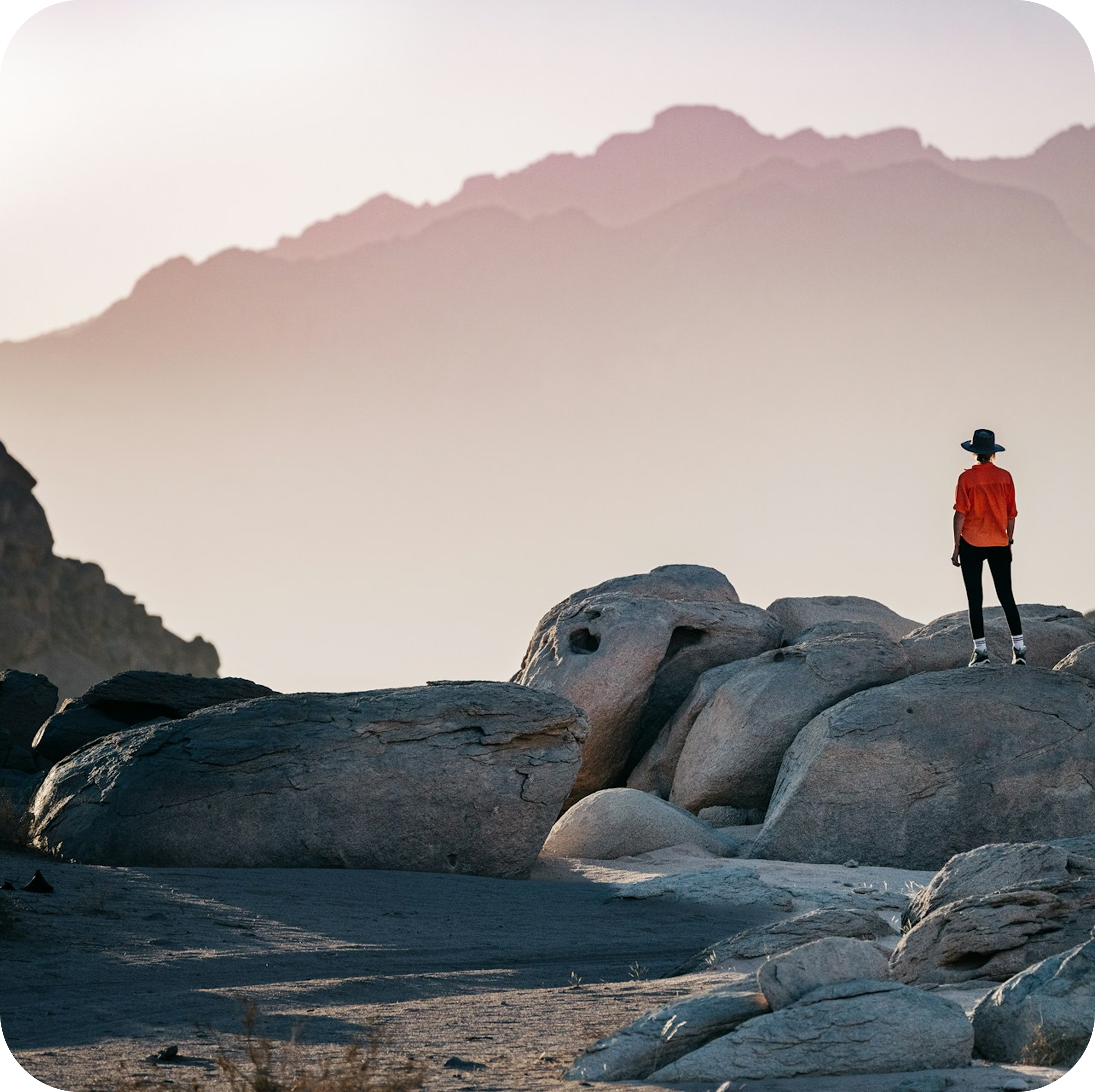 Person in orange shirt standing on rocky outcrop overlooking mountain landscape at dusk